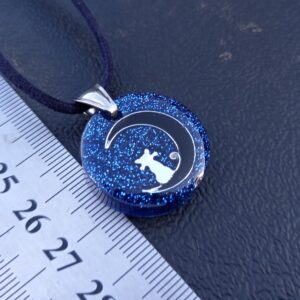 A cat sitting on a crescent moon in the glittery blue night sky. Stainless Steel. Navy faux necklace.